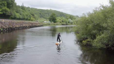 Aerial-overtakes-man-on-paddleboard-enjoying-calm-rural-day-on-river
