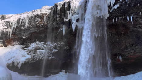 Spektakulärer-Seljalandsfoss-wasserfall-Im-Winterisland,-Niedriger-Winkelneigungsblick-Auf-Die-Kaskade-Der-Fälle-Und-Wasser,-Das-Von-Einer-Hohen-Felsigen-Schneebedeckten-Klippe-Herunterfließt