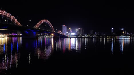 TimeLapse-Night-Panoramic-View-of-Hàn-River-in-Da-Nang-City-Vietnam,-illuminated-Dragon-Bridge,-Colorful-Reflection-of-Lights-on-Water-and-Boats-Sailing-Passing,-Downtown-Buildings-in-Horizon