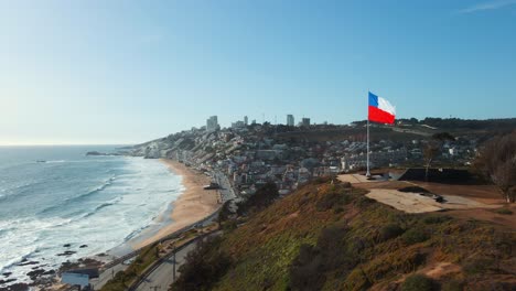 Aerial-view-passing-close-to-republic-of-Chile-flag-flying-on-Reñaca-hilltop-revealing-downtown-city-coastal-buildings