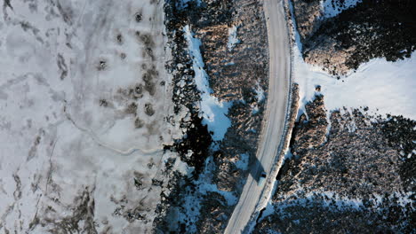 Aerial-Top-Down-View-Tracking-Van-Car-Driving-Moving-in-Winter-on-Curved-Road-Through-Wild-Snowy-Rocky-Lands-and-Frozen-Wetlands-in-Búlandstindur-Mountain-Area,-Iceland