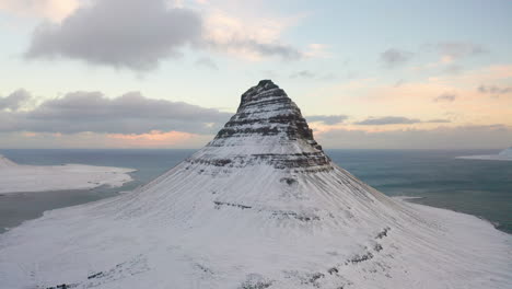 Aerial-Orbit-View-of-Kirkjufell-Mountain-Iceland-in-Winter,-Covered-with-Snow-Sharpened-Rocky-Peak-at-the-Top,-Long-High-Curved-Sides-and-White-Snowy-Base-Lands,-Cloudy-Sky-at-Sunset-in-Background