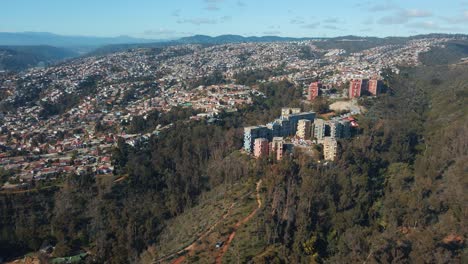 Aerial-orbit-of-colorful-buildings-in-hillside-Quinta-Vergara-Park-covered-in-forest,-Viña-del-Mar-city-in-background,-Chile