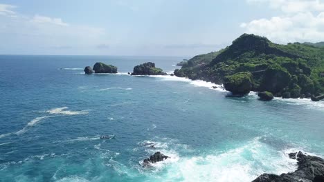 Aerial-flyover-bay-with-blue-water-and-ocean-waves-crashing-against-cliffs-and-rocks-in-sunlight---Timang-Island,Yogyakarta,Indonesia