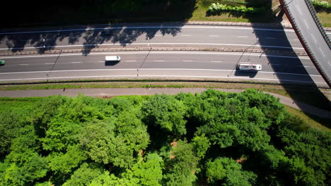 Top-down-aerial-of-cars-and-trucks-driving-on-highway-during-sunny-day-beside-trees