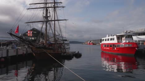 Slow-pan-up-out-of-Constitution-Dock-area-over-old-wooden-sailing-boat-and-spirit-of-Hobart-red-boat-with-calm-water-on-clear-winter-day-as-people-walk-past,-Hobart,-Tasmania