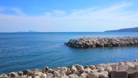 Rock-Breakwater-Surrounded-By-Calm-Blue-Waters-With-Capri-Island-In-The-Distance-At-Naples,-Italy