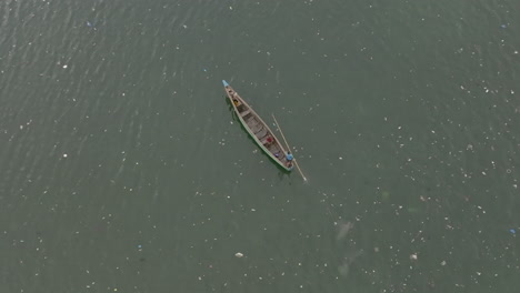 Aerial-footage-of-a-long-boat-slowly-traveling-through-the-water-with-trash-all-around-it