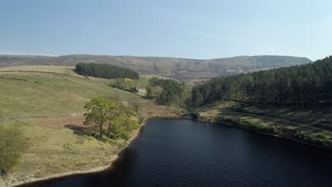 Aerial-shot-panning-left-over-Kinder-Reservoir-water-close-to-trees-revealing-a-National-Trust-estate-in-the-distance-in-the-Kinder-Scout-Valley