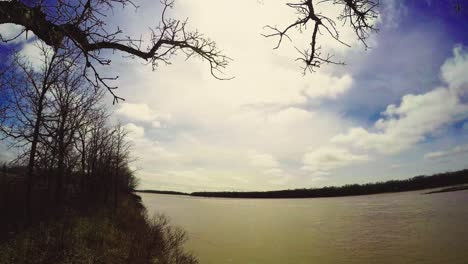 Early-spring-riverside-timelapse-of-two-levels-of-clouds-on-sunny-day-with-trees-and-branches-at-riverside