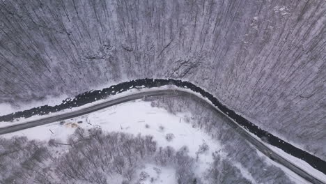 [Aerial-shot]-Car-passing-by-on-road-near-a-river-that-separates-the-forest-with-trees-covered-by-snow