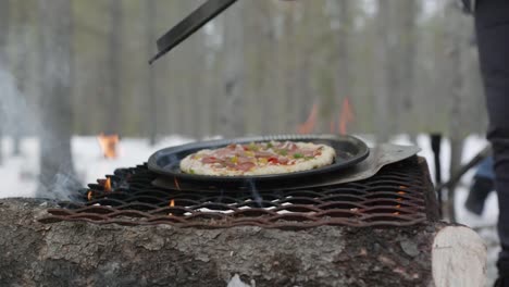 Removing-a-steel-lid-to-show-a-pizza-being-cooked-on-a-steel-grate-over-an-open-wood-fire