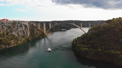 Sailing-boat-driving-under-car-bridge-in-fjord-captured-with-drone