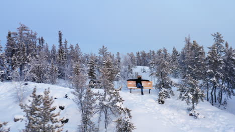 Man-sits-on-a-bench-in-remote-snow-wilderness,-Aerial-close-to-far-out