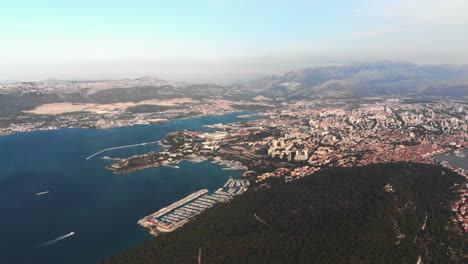 Drone-slowly-turning-from-the-big-city-of-Split,-over-the-harbor-ans-stadion-to-the-Adriatic-sea-with-a-red-ferry