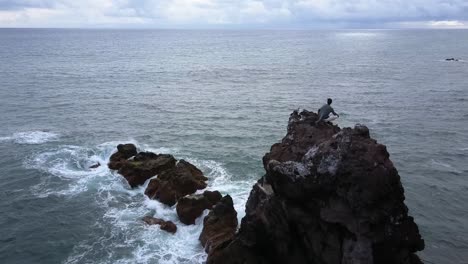 Madeira-Insel,-Portugal---Mann-Sitzt-Auf-Einer-Klippe-Und-Beobachtet-Die-Meereswellen,-Die-Auf-Die-Felsen-Spritzen---Luftdrohnenaufnahme