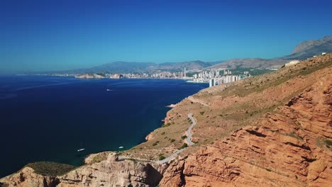 Aerial-view-of-empty-curve-and-turn-on-road-on-mountain-surrounded-by-water-with-buildings-on-cliff-with-a-yacht-sailing-in-the-calm-ocean-on-a-clear-sky-day-in-Benidorm-city-of-Spain