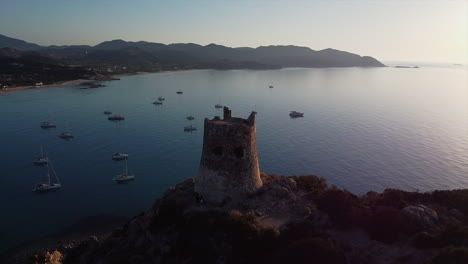 Establisher-shot-of-Torre-di-Porto-Giunco-tower-surrounded-by-rocks-and-shrubs-in-Sardinia-in-Italy-during-sunset-evening