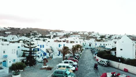 Aerial-view-of-three-people-travelling-round-the-city-on-bike-in-the-beautiful-empty-lanes-of-Mykonos-with-cars-parked-outside-traditional-white-houses