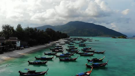 Aerial-view-of-traditional-boats-parked-at-coast-of-sandy-beach-with-sparkling-clear-water-of-sea-surrounded-by-mountains-on-a-cloudy-day-in-Thailand