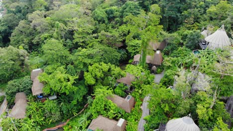 Top-down-view-of-luxurious-resort-over-hill-surrounded-by-greenery-with-small-huts-and-houses-spread-across-the-city-with-calm-sea-and-mountains-in-Thailand