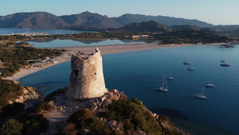 Panoramic-circular-shot-of-beautiful-Torre-di-Porto-Giunco-tower-surrounded-by-mountain,-beach-and-sea-with-yacht-sailing-during-sunset