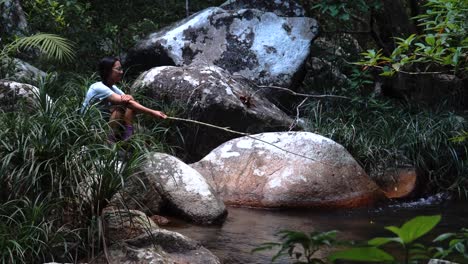 Asian-woman-sitting-on-rock-and-fishing-with-wooden-stick-in-jungle-stream