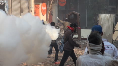 Ein-Maskierter-Demonstrant-Läuft-Mit-Einem-Rauchenden-Tränengaskanister-Und-Wirft-Ihn-Während-Der-Zusammenstöße-In-Der-ägyptischen-Revolution-Auf-Einer-Straße-In-Kairo-In-Richtung-Der-Sicherheitskräfte-Zurück