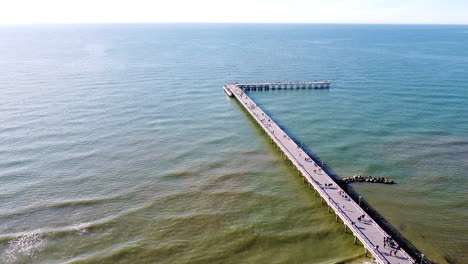 People-walking-on-Palanga-bridge-on-sunny-day-with-calm-sea,-aerial-ascend-view