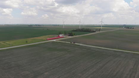 Tractor-Tirando-De-Equipo-Agrícola-En-Granja-Camino-De-Tierra-Campos-Turbina-Eólica-Iowa-America-Drone-Aéreo