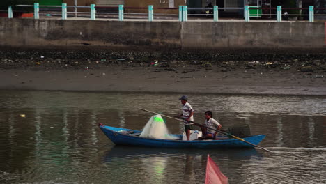 Dos-Pescadores-Vietnamitas-Remando-En-Un-Bote-De-Pesca-En-El-Río-Contaminado-De-La-Ciudad-En-Vietnam
