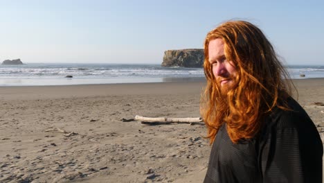 A-man-with-long-red-hair-looks-out-over-a-beach-in-Oregon-contemplating-calmly