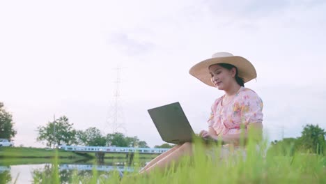 Schöne-Asiatische-Frau-Mit-Hut,-Die-Auf-Dem-Rasen-Sitzt-Und-Am-Computer-Arbeitet-Und-Sich-Erfolgreich-Fühlt