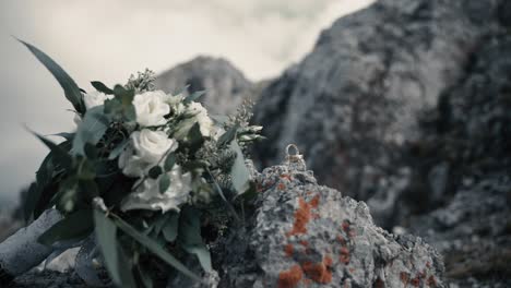 Brautblumenstrauß-Mit-Weißen-Rosen-Und-Goldenen-Ringen,-Die-Auf-Einem-Felsen-Auf-Einem-Berggipfel-Der-Alpen-Liegen,-Während-Der-Wind-Die-Blätter-Weht