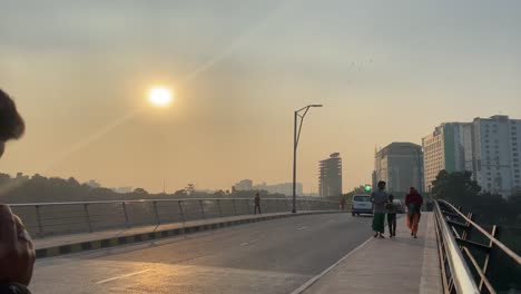 Static-view-of-sunset-from-a-bridge-over-a-canal-with-people-walking-by-on-the-pavement,-cars-and-vans-passing-by-in-Dhaka,-Bangladesh