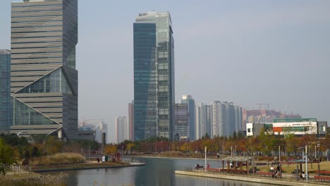 Los-Edificios-De-Oficinas-De-La-Torre-G-Y-La-Torre-Ibs-En-El-Parque-Central-De-Incheon-Songdo-En-Otoño-Con-Gente-Caminando-Cerca-Del-Lago---Estática-Aérea