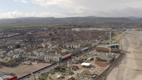 An-aerial-view-of-Rhyl-promenade-and-seafront-on-a-cloudy-day,-flying-away-from-the-town-centre-over-the-beach,-North-Wales,-UK