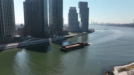 An-aerial-view-of-a-barge-sailing-down-Newtown-Creek-with-new-high-rise-apartment-buildings-in-Brooklyn,-NY-in-the-background-on-a-sunny-day