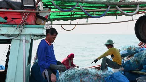 Un-Pescador-Reparando-Redes-Mientras-Fuma-Mientras-Se-Saca-El-Cigarrillo-De-La-Boca-Mientras-Otros-También-Trabajan-En-El-Fondo,-Muelle-De-Pesca-De-Pattaya,-Chonburi,-Tailandia