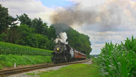 A-Front-View-of-an-Antique-Steam-Passenger-Train-Pulling-out-of-a-Stop-and-Traveling-Thru-Farmlands-and-Corn-Fields-Blowing-Black-Smoke-on-a-Sunny-Summer-Day