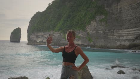 Charming-blond-woman-taking-selfies-at-tropical-beach-with-tall-cliffs-in-background,-joyful-vacation-moment