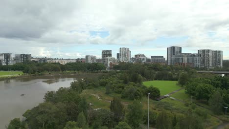 Aerial-fly-over-a-beautiful-green-park-and-river-with-cityscape-in-the-suburb-of-Sydney,-Australia