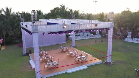 aerial-footage-of-spacious-event-location-by-the-water,-pastel-themed-event-set-up,-symmetrical-chair-set-up-on-the-large-evergreen-lawns,-colorful-vibes,-bright-outdoor-day,-event-space-for-several