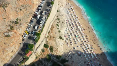 aerial-top-down-view-of-traffic-on-a-coastal-road-alongside-a-cliff-with-the-Mediterranean-Sea-below-with-dark-blue-tropical-water-and-European-tourists-relaxing-at-Kaputas-Beach-in-Kas-Turkey