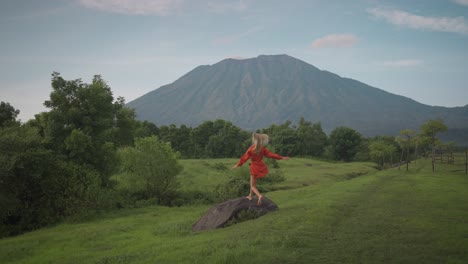 Blond-woman-in-orange-dress-runs-barefoot-through-Savana-Tianyar-grass-field-onto-rock-boulder,-turning-around,-view-of-Mount-Agung,-travel-girl-concept