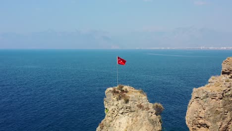Drone-Aéreo-Pasando-Lentamente-La-Bandera-Turca-De-Pie-Sobre-Una-Roca-En-Medio-Del-Mar-Mediterráneo-En-Antalya-En-Un-Día-Soleado-De-Verano