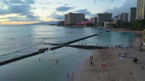 Turistas-Disfrutando-De-La-Playa-De-Waikiki-En-Honolulu,-Hawaii-Con-Surfistas-En-Las-Olas-Del-Océano-Fuera-De-Un-Rompeolas-Donde-La-Gente-Nada