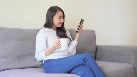 Asian-woman-with-cup-of-tea-sitting-on-a-couch-and-watching-phone-with-happy-face-expression