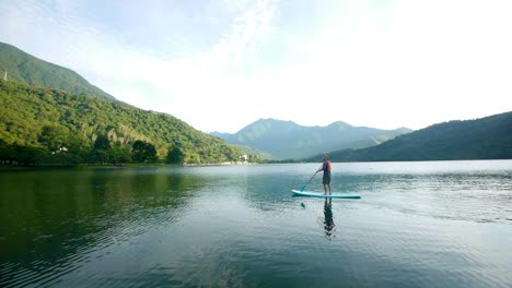 Asian-man-on-stand-up-paddle-board-moving-through-water-at-fast-pace-over-lake-surrounded-by-mountains,-filmed-as-wide-shot-during-early-morning