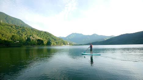 Asiatischer-Mann-Auf-Stand-Up-Paddle-Board,-Der-Sich-Schnell-Durch-Wasser-über-Einen-Von-Bergen-Umgebenen-See-Bewegt,-Gefilmt-Als-Totalaufnahme-Am-Frühen-Morgen